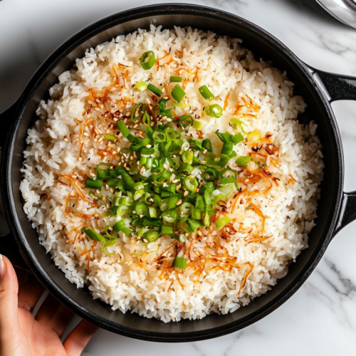 This image shows a large bowl of perfectly cooked Asian rice, garnished with green onions and served hot. The fluffy jasmine rice, mixed with soy sauce and sesame oil, is complemented by the fresh green onions and optional peas, ready to be enjoyed as a flavorful side dish.