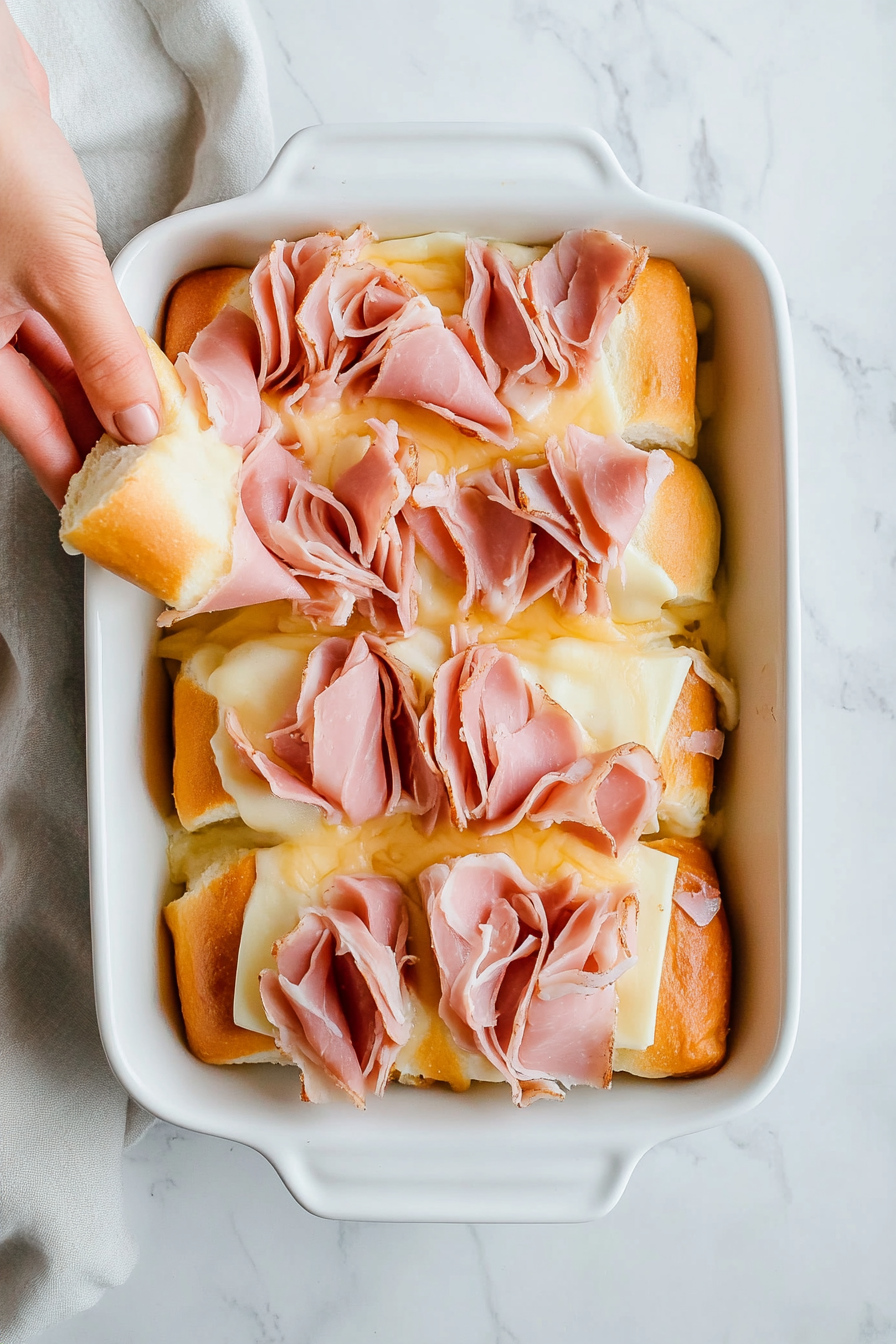 A white 9x13 baking dish sits on a white marble countertop, filled with the bottom halves of soft Hawaiian slider rolls neatly arranged side by side. This step forms the base for the funeral sandwiches, preparing them for layering with cheese and ham. A hand is seen placing the last roll, completing the foundation layer.