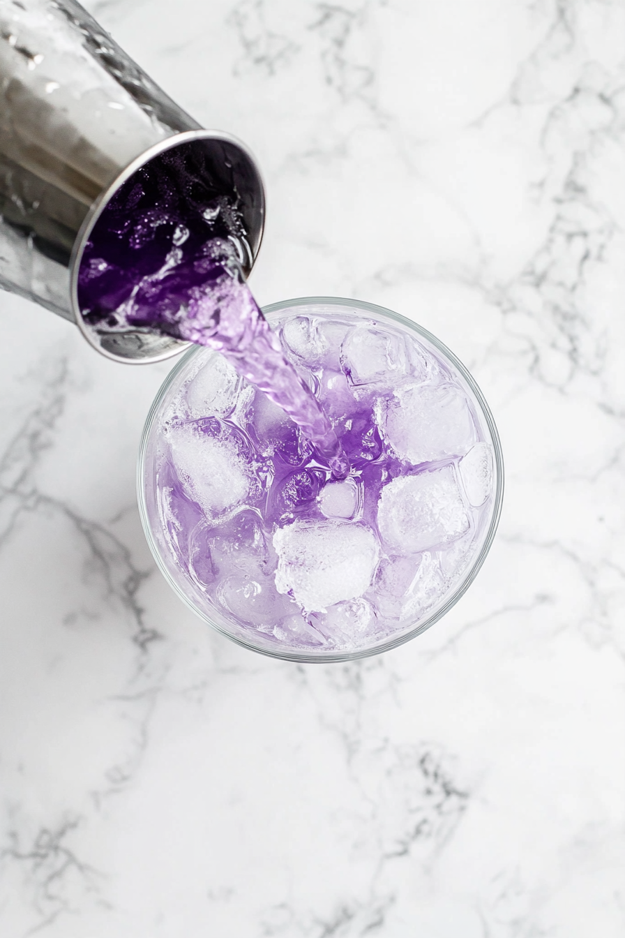 This image shows the chilled Transfusion cocktail, made with vodka, grape juice, and lime, being poured into a glass filled with ice cubes