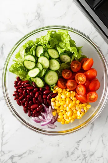 Top-down view of a large bowl filled with fresh lettuce, kidney beans, sliced cucumber, corn, halved cherry tomatoes, and thinly sliced red onion. The colorful ingredients are layered on a white marble cooktop.