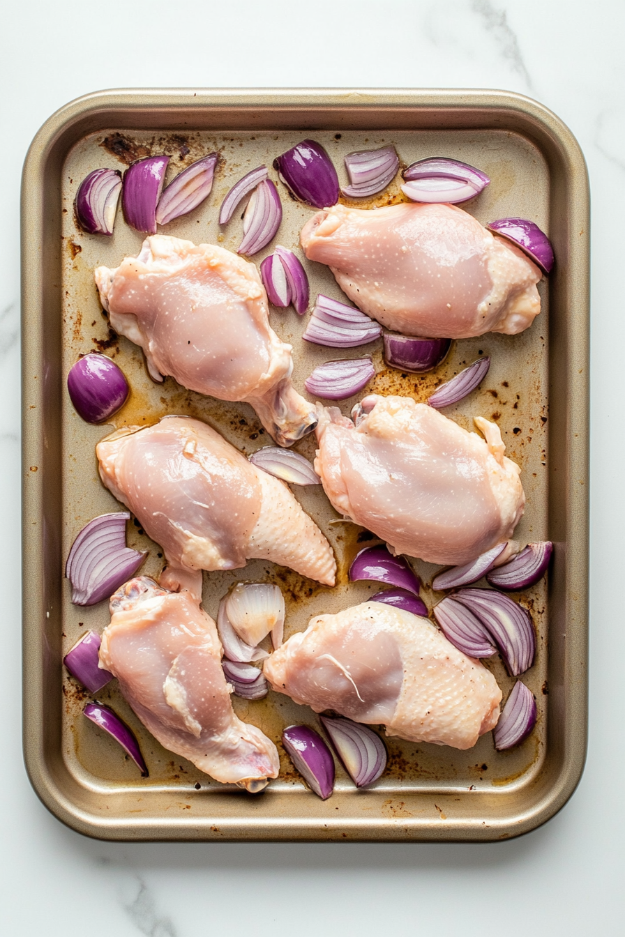 This image shows the marinated chicken legs and thighs arranged on a baking sheet, nestled between halved shallots, ready to go into the oven for roasting.