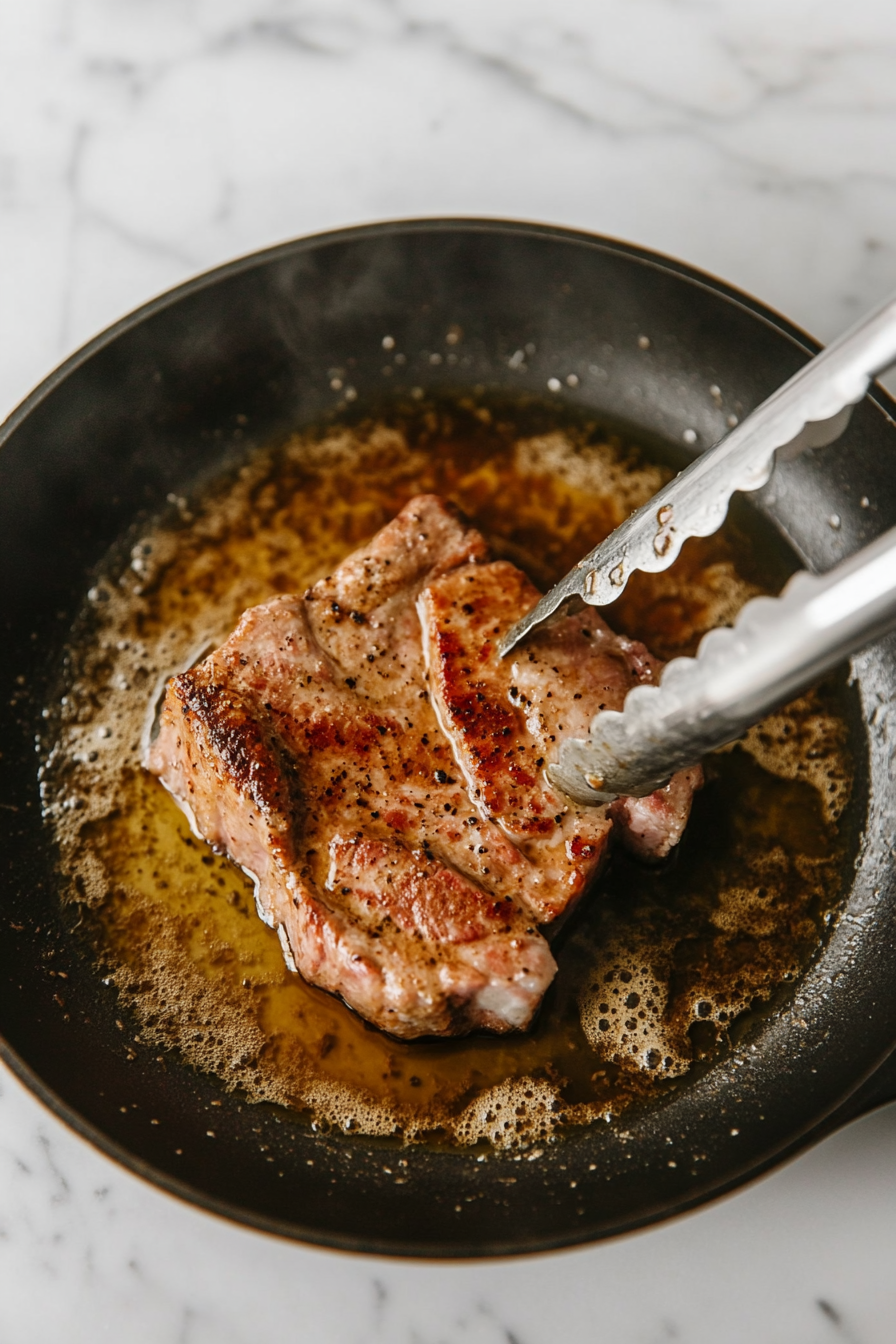 This image shows a pork ribeye roast being seared in a large skillet, turning golden brown on all sides as it cooks over medium-high heat to develop a flavorful crust.