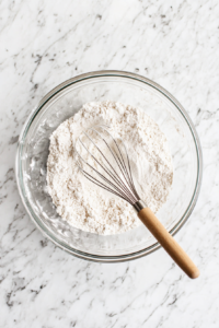 This image shows uncooked quick-cooking grits being slowly poured into the saucepan of hot broth and milk, beginning the cooking process for the creamy Gouda grits.