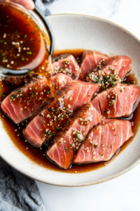 This image shows yellowfin tuna steaks being marinated in a dish filled with a mixture of coconut aminos, honey, ginger, and garlic to infuse flavor.