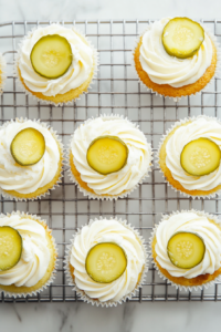 This image shows freshly baked pickle cupcakes being frosted with a creamy bourbon-infused frosting and garnished with tangy pickle slices.