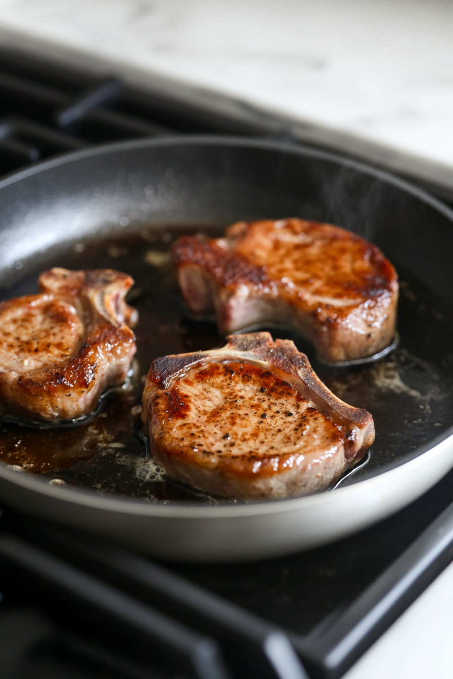 This image shows pork chops sizzling in a skillet, achieving a golden-brown crust before being transferred to the oven for baking.