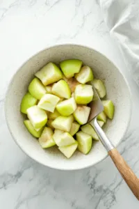 A medium bowl filled with chopped Granny Smith apples being coated with 2 tablespoons of sugar and 1 teaspoon of cinnamon. A spatula is stirring the mixture, highlighting the contrast between the vibrant green apples and the sugar-cinnamon coating