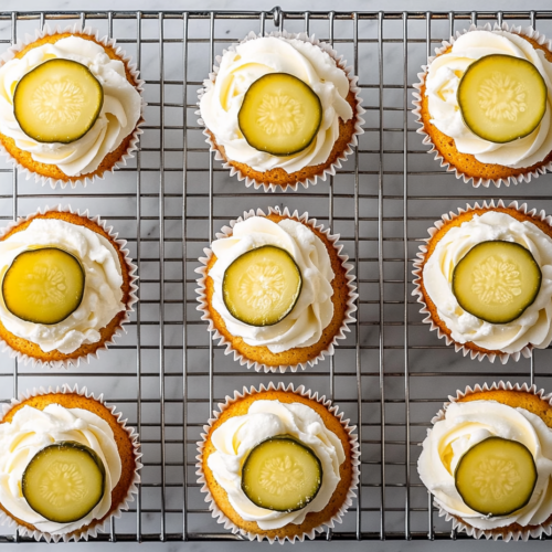 This image shows several pickle cupcakes topped with a smooth bourbon cream cheese frosting and garnished with small pickle slices, offering a visually intriguing and savory twist on a classic dessert.