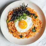 This image shows a vibrant serving of kimchi fried rice on a plate, topped with crispy bacon, a perfectly cooked sunny-side-up egg, and garnished with sesame seeds and green onions for a delicious Korean comfort meal.