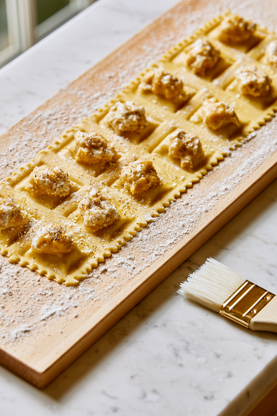 Lightly flour a ravioli maker and place a pasta sheet over it, gently pressing to create pockets. Add about 1 tablespoon of pumpkin filling into each pocket, brush the edges with water, and cover with another pasta sheet
