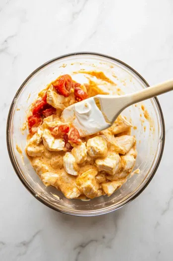 A mixing bowl on the white marble cooktop is filled with diced chicken, 1 cup of sour cream, lime juice, diced tomatoes (14.5 oz), and ½ packet of taco seasoning. A rubber spatula is used to combine the ingredients, emphasizing the creamy consistency of the mixture.