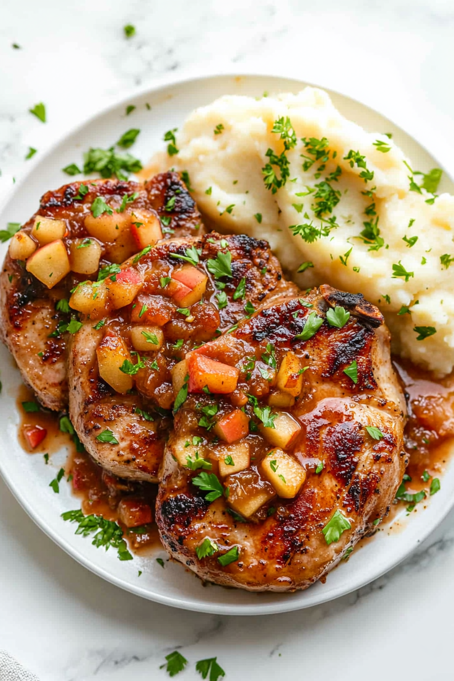 This image shows the fully cooked pork chops plated with a generous serving of apple chutney on top, garnished with fresh parsley, and served with mashed potatoes.