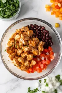 A mixing bowl on a white marble cooktop contains 1 lb of cooked and diced chicken tenderloins. Nearby, a can of black beans (14.5 oz) is being drained and rinsed, alongside diced white onion and red bell pepper. The scene highlights the fresh ingredients ready for the casserole.