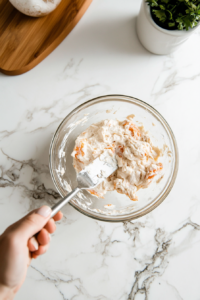 This image depicts a spoon transferring the prepared Cold Crab Dip from the mixing bowl into a small serving dish, ready to be chilled in the refrigerator before serving to guests.