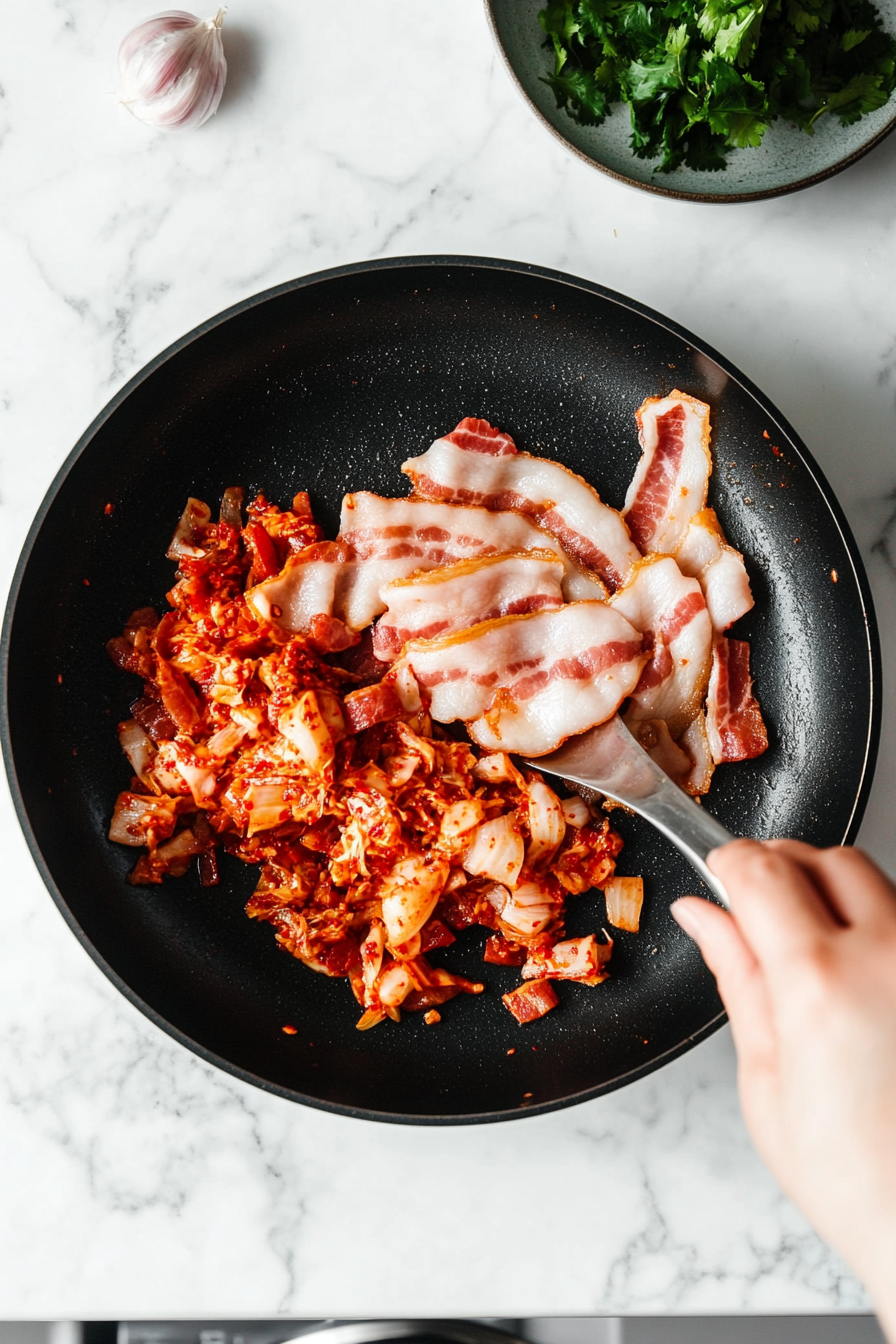 This image shows kimchi and minced garlic being stirred into the pan with the cooked bacon, creating a blend of spicy, tangy, and savory flavors for the fried rice.