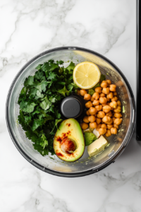 This image shows all the ingredients, including ripe avocado, chickpeas, tahini, lemon juice, olive oil, garlic, and cilantro, being placed into a food processor for blending into avocado hummus