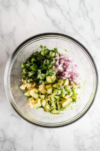 This image shows diced avocado, shredded chicken, red onions, and lime juice being added to a large mixing bowl, preparing to create a flavorful avocado chicken salad.