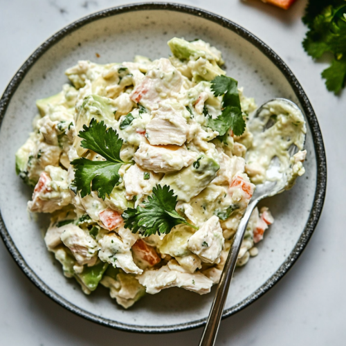 This image shows a creamy avocado chicken salad served on a white plate, with fresh lime wedges and cilantro on the side, highlighting its vibrant colors and rich texture, perfect for a light and healthy meal.