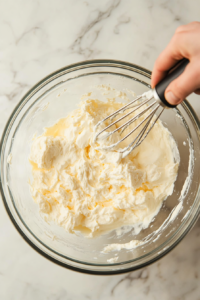 This image shows a large mixing bowl with cream cheese, eggs, and sugar being whipped together to create a smooth and creamy cheesecake filling, which will be poured over the graham cracker crust for the Crème Brûlée cheesecake.