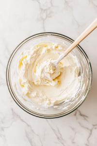 This image shows softened cream cheese, sugar, and whipped topping being mixed together in a large bowl, creating a smooth and creamy filling for the cheesecake.