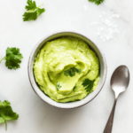 This image shows a bowl of creamy avocado crema with a spoon, garnished with fresh cilantro and lime wedges, ready to be drizzled over tacos, nachos, or grilled meats for a refreshing and flavorful addition.