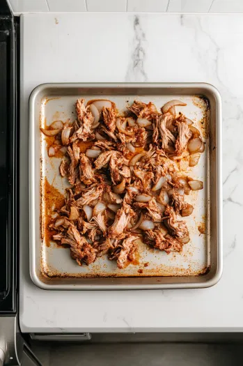 Top-down view over a white marble cooktop with a baking sheet holding shredded meat and onions tossed with additional taco seasoning. The oven is set to 425°F, with the baking sheet ready to be placed inside. The scene captures the crisping process, highlighting the transformation of the meat as it caramelizes for a crispy texture.