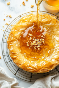 This image shows warm honey syrup being poured over the edges of the baked baklava cheesecake, soaking into the crispy phyllo layers for added sweetness and texture.