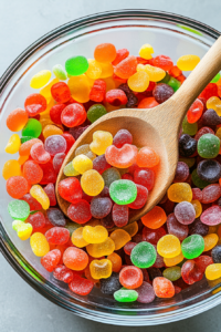 This image shows a large wooden spoon carefully stirring the mix of gummy candies in the bowl. The candy salad is a rainbow of colors and shapes, featuring gummy bears, worms, peach rings, and more, blending together for a fun, chewy treat. The spoon is ensuring even distribution of all the candies.