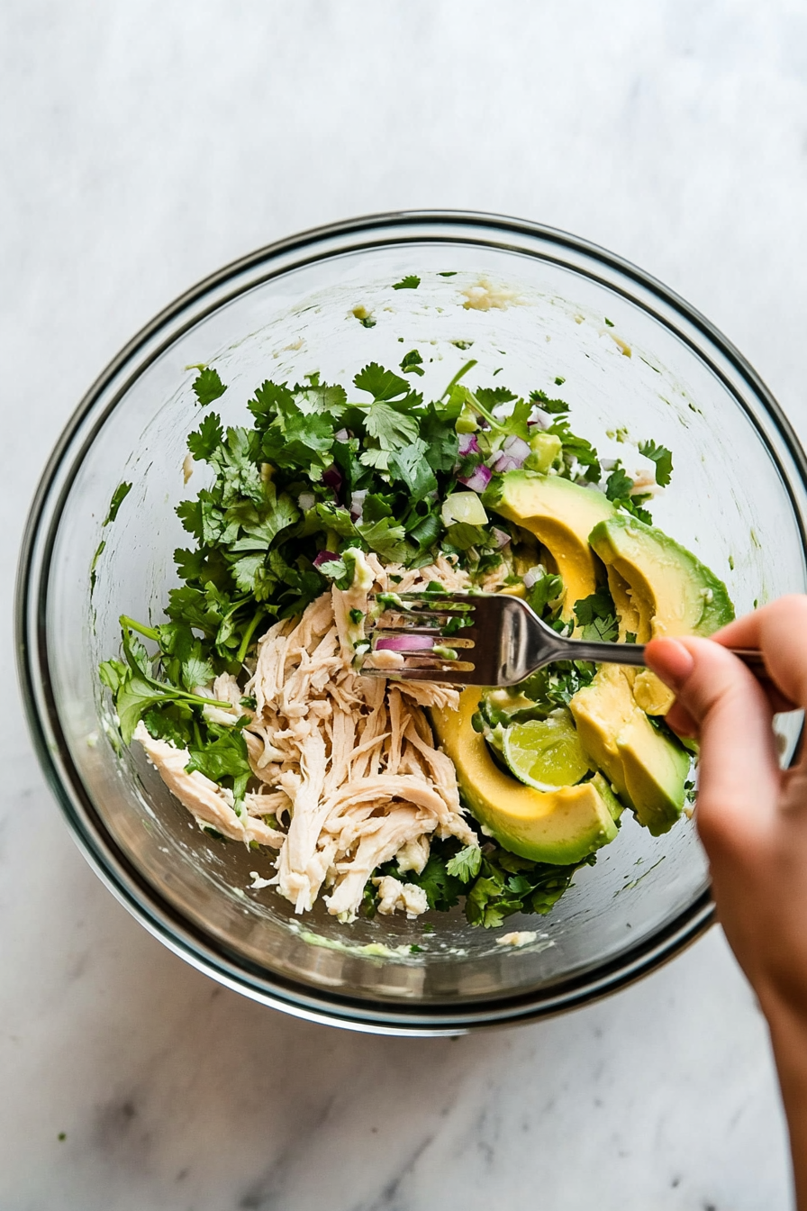 This image shows a fork mashing avocado in a mixing bowl while combining it with shredded chicken, red onion, cilantro, and lime juice, blending into a smooth, creamy chicken salad.