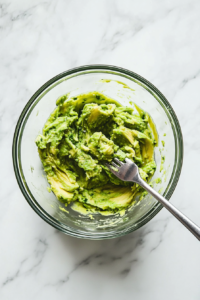 This image shows a mixing bowl with ripe avocado being mashed together with lemon juice, garlic powder, and salt, creating a creamy topping for the avocado burgers.