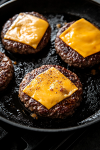 This image shows slices of cheddar cheese melting on top of freshly cooked beef patties in a skillet, preparing them for the avocado burger assembly.