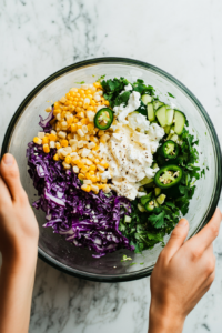 This image shows a large bowl with shredded cabbage, charred corn, jalapeños, and green bell peppers being mixed together. The slaw is colorful and ready to be coated with the zesty lime dressing, combining fresh ingredients with bold flavors.