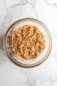 This image shows a large mixing bowl filled with crushed graham crackers, granulated sugar, and melted butter, being mixed on a clean kitchen countertop. The crumbs are evenly coated and ready to be pressed into the base of a 9-inch springform pan lined with parchment paper to create the crust for the Crème Brûlée cheesecake.