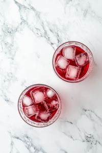 This image shows vibrant red raspberry syrup being carefully poured into two large drinking glasses, creating the sweet-tart base for the raspberry Italian soda.