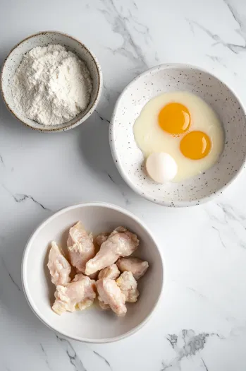 A cutting board on a white marble cooktop where a large boneless, skinless chicken breast is being cut into bite-sized pieces. A sharp knife is nearby, highlighting the preparation of the chicken for frying.