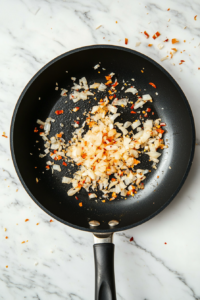 This image shows garlic, minced ginger, bird eye chilies, and chopped yellow onions sizzling in ghee, releasing aromatic flavors that enhance the Palak Sauce.