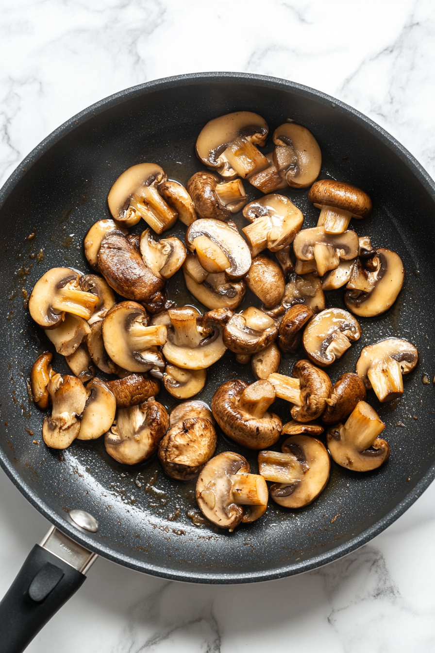 This image shows garlic being gently sautéed in oil, followed by baby bella mushrooms browning in the pan, releasing a rich, earthy aroma that forms the base for the creamy Mushroom Alfredo sauce.