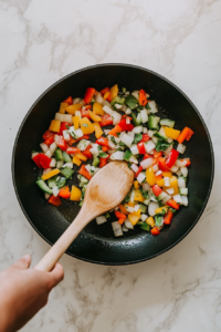 This image shows a large skillet with diced yellow onion, red bell pepper, yellow bell pepper, and poblano chili sautéing in olive oil over medium heat, creating a colorful and aromatic base for the Southwest Chicken Bowl.