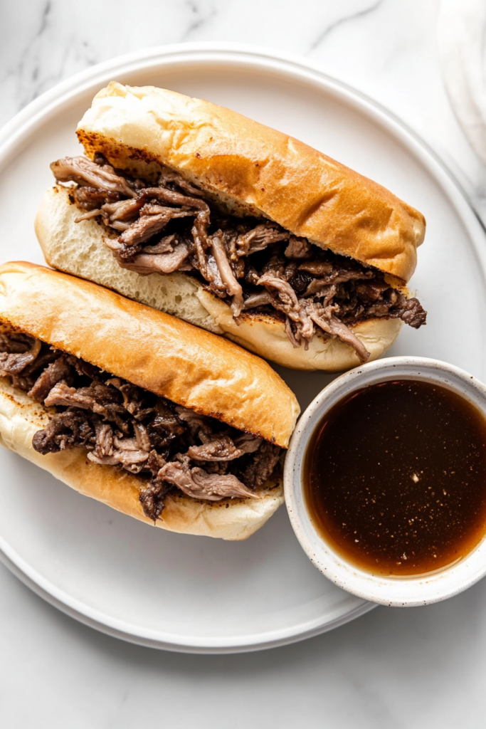 This image shows French dip sandwiches plated alongside a bowl of warm, reduced au jus, perfect for dipping the sandwich into the flavorful broth.