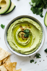 This image shows a bowl of freshly blended avocado hummus, garnished with a drizzle of olive oil and served alongside pita chips and sliced vegetables for dipping.