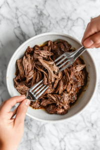 This image shows the tender chuck roast being shredded into juicy pieces with two forks in a bowl, ready to be assembled into sandwiches.