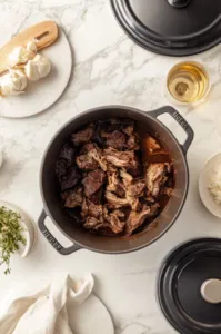 Top-down view over a white marble cooktop with the Dutch oven removed from the oven. The lid is lifted, revealing tender, cooked chuck roast. Two forks are shredding the meat into large chunks on a clean plate, showcasing the juiciness and ease of shredding.