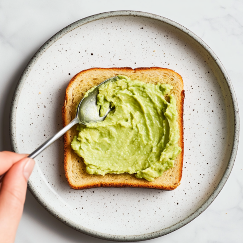 This image shows a perfectly toasted slice of bread topped with creamy mashed avocado, sprinkled with sea salt and chili flakes, making a quick and nutritious avocado toast snack.