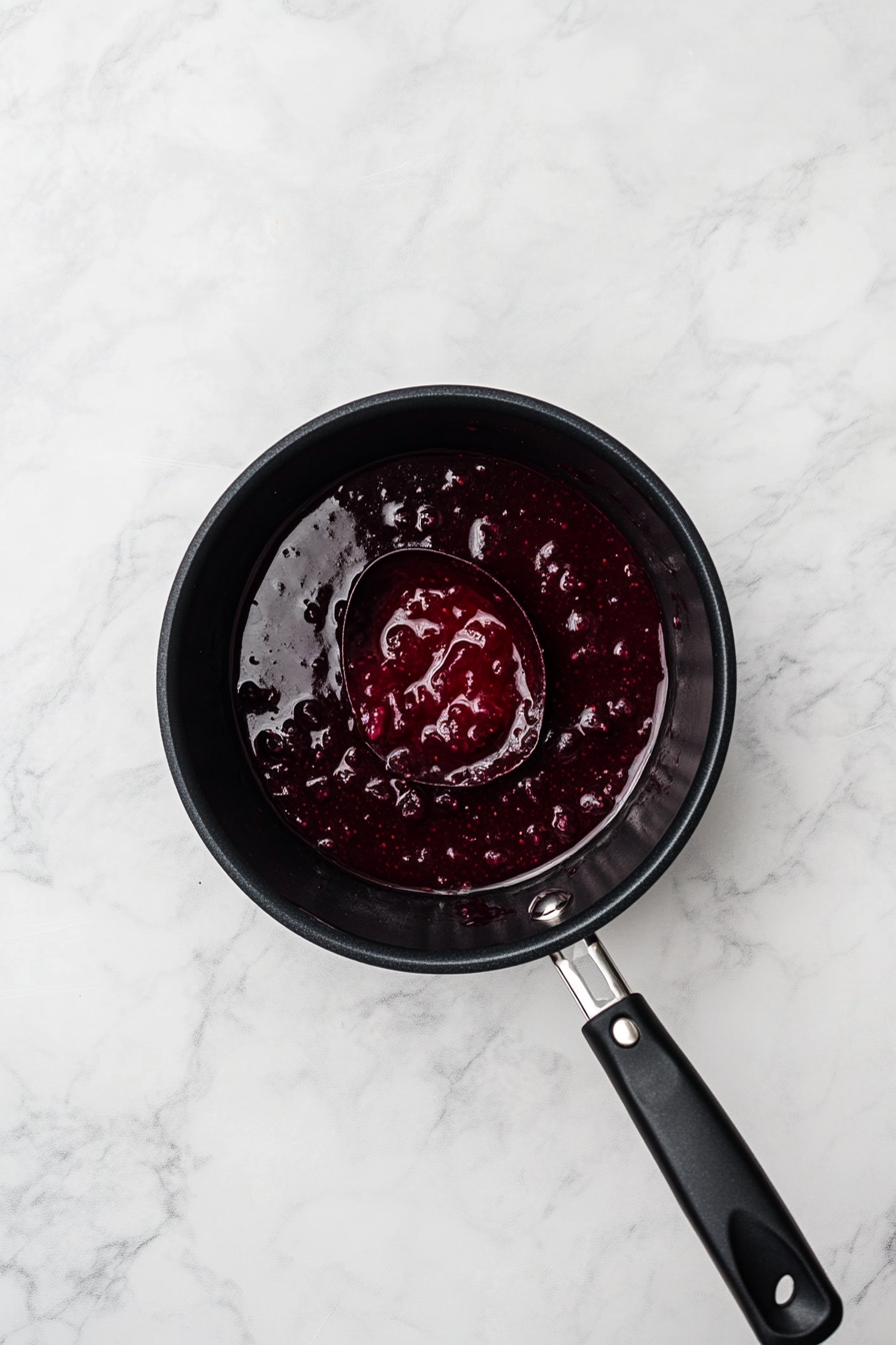 This image shows huckleberries, water, cornstarch, and sugar being stirred in a saucepan over medium heat, cooking into a thick and tangy topping for the cheesecake.