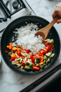 This image shows minced garlic being added to the skillet along with fluffy basmati rice, stirring to coat the grains with the flavorful sautéed vegetables in preparation for the Southwest Chicken Bowl.