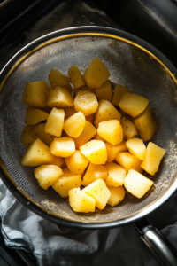 This image shows boiled potato cubes being drained in a strainer and gently patted dry with a dish towel to eliminate excess moisture for crispier hashbrowns.