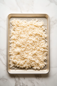 This image shows fluffed rice spread evenly on a large baking sheet, cooling completely to ensure the rice will achieve a perfect crispy texture.