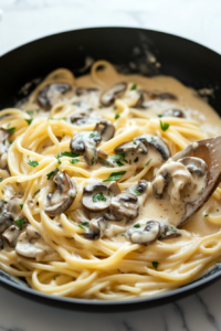 This image shows cooked pasta being tossed into the rich Alfredo sauce with mushrooms, blending the flavors together and ensuring the pasta is fully coated in the creamy mixture.