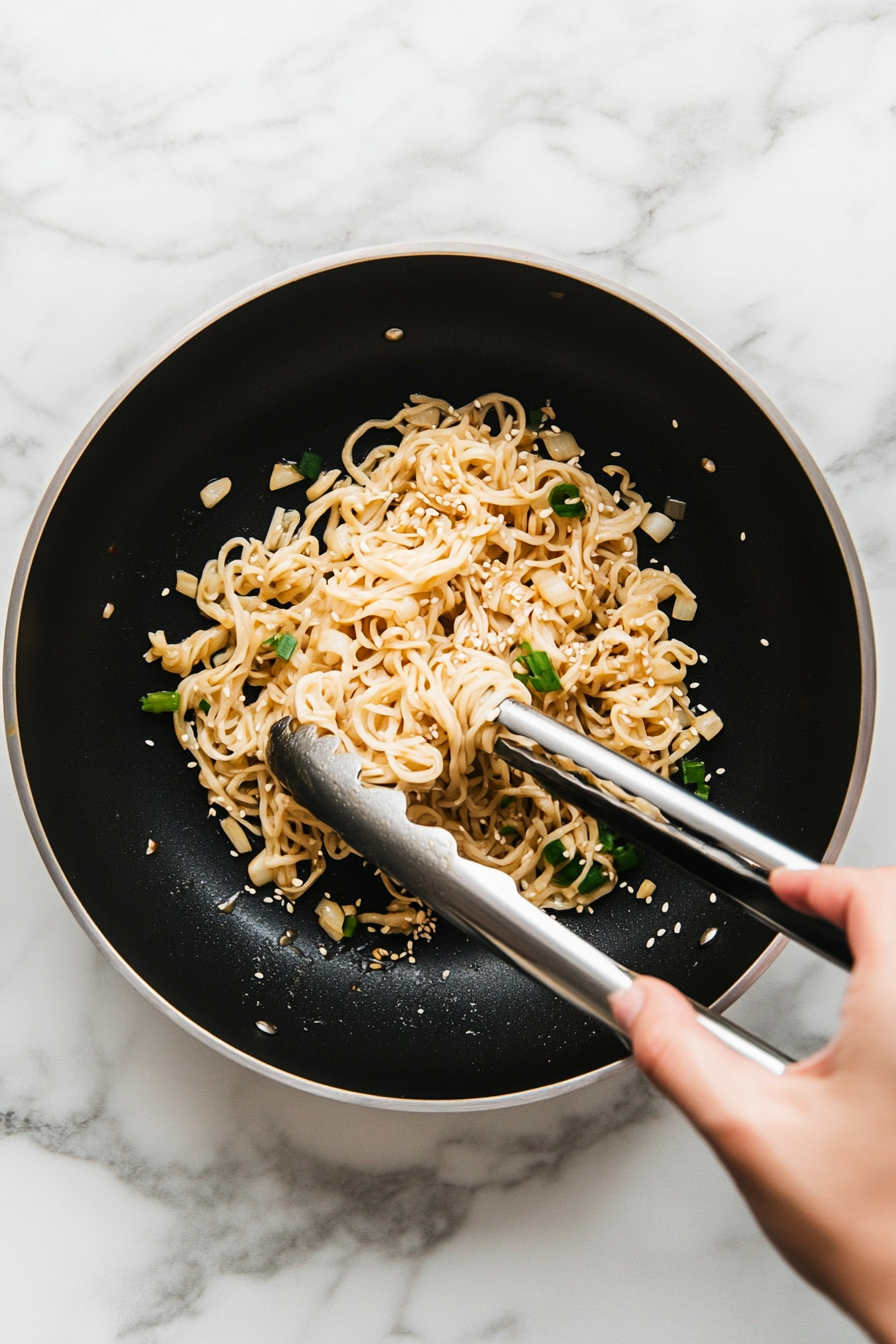 This image shows cooked noodles being tossed in a pan with homemade teriyaki sauce, garlic, and oil, ensuring they are fully coated before serving.