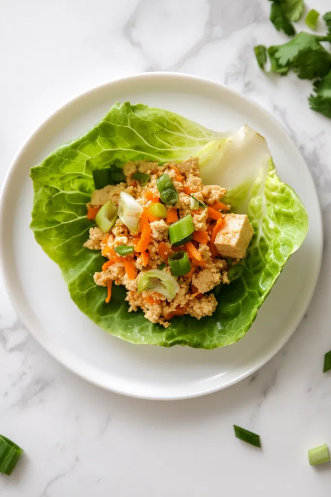 This image shows the finished vegan lettuce wraps filled with tofu, mushrooms, water chestnuts, and hoisin sauce, topped with grated carrots and green onions, and served on a platter.